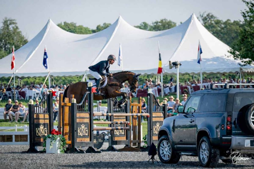 Horse jumping in stadium ring with white tent and spectator in background