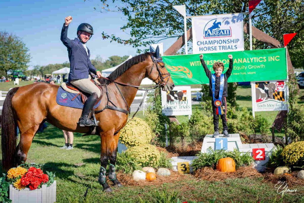 Man on horse raising arm, boy with ribbon around neck raising arms while standing on podium.
